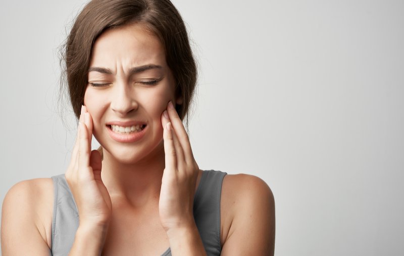 a young woman wearing a gray tank top and holding both sides of her jaw in pain